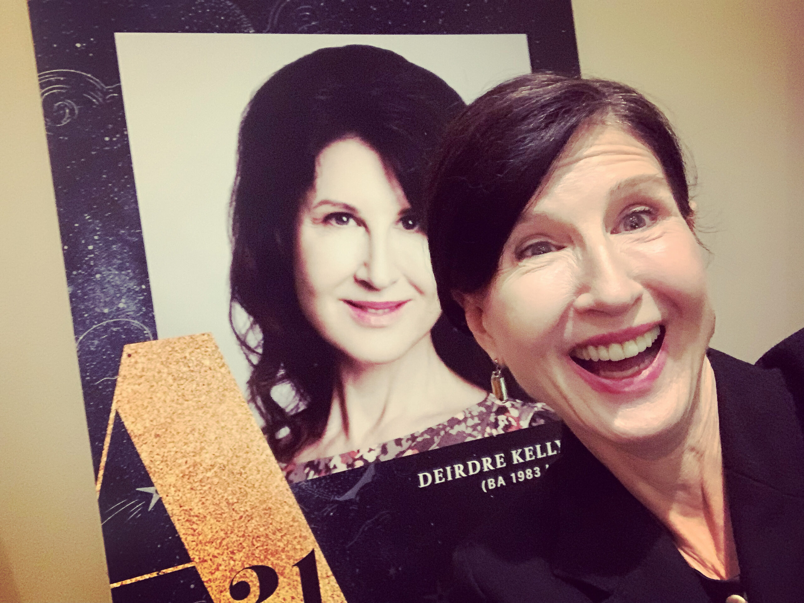 A smiling woman takes a selfie in front of a poster featuring a similar woman. The poster includes gold and black decorative elements, conveying a celebratory tone.
