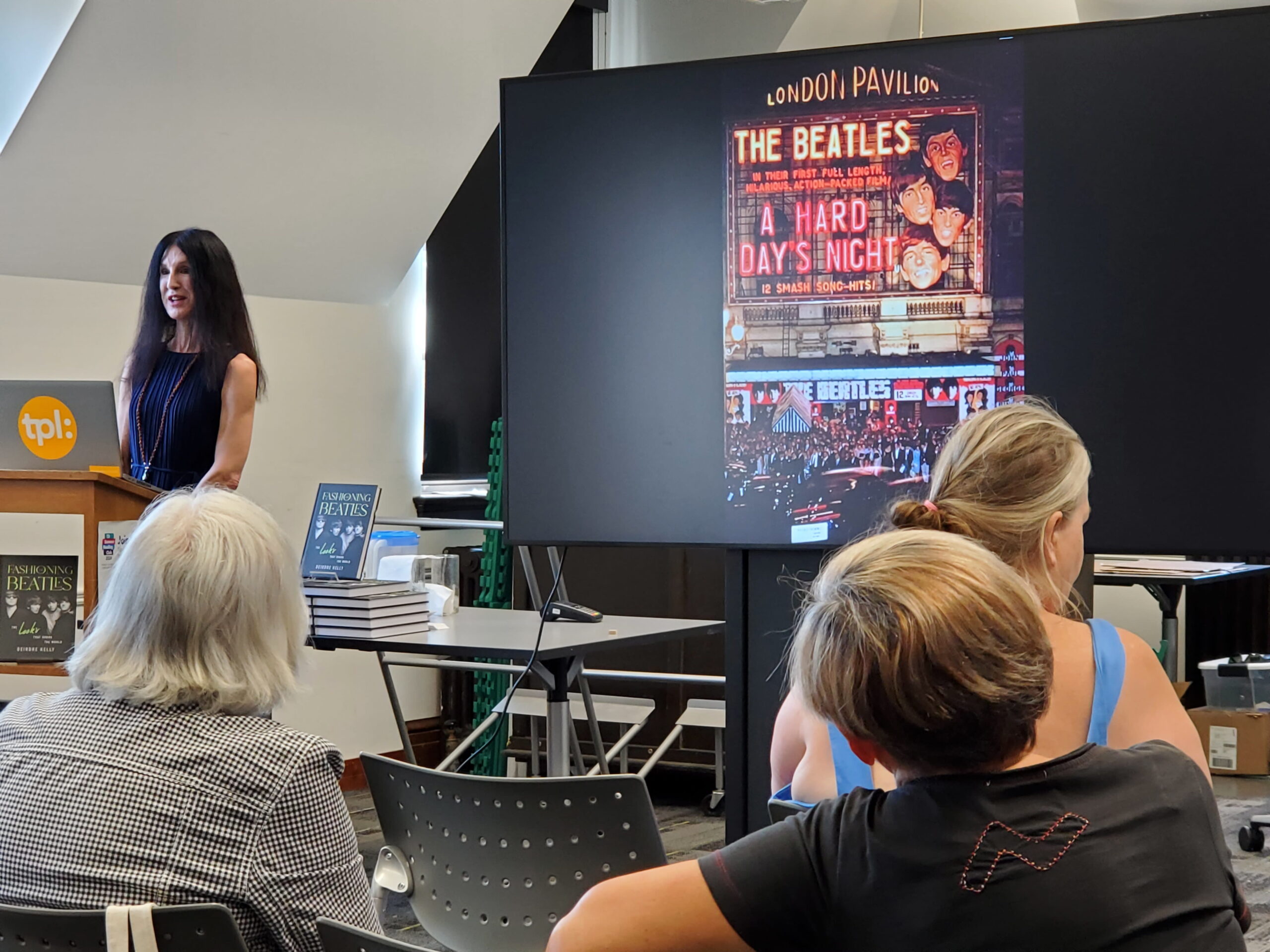 A woman stands at a podium with a screen displaying "The Beatles: A Hard Day's Night" poster. Three attendees sit facing the presentation. The room feels educational and focused.