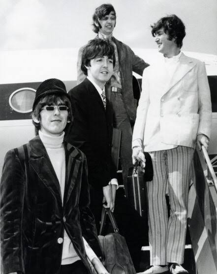 The Beatles disembark from a plane, showcasing their iconic fashion. Paul and George smile at the camera, while John and Ringo carry briefcases.