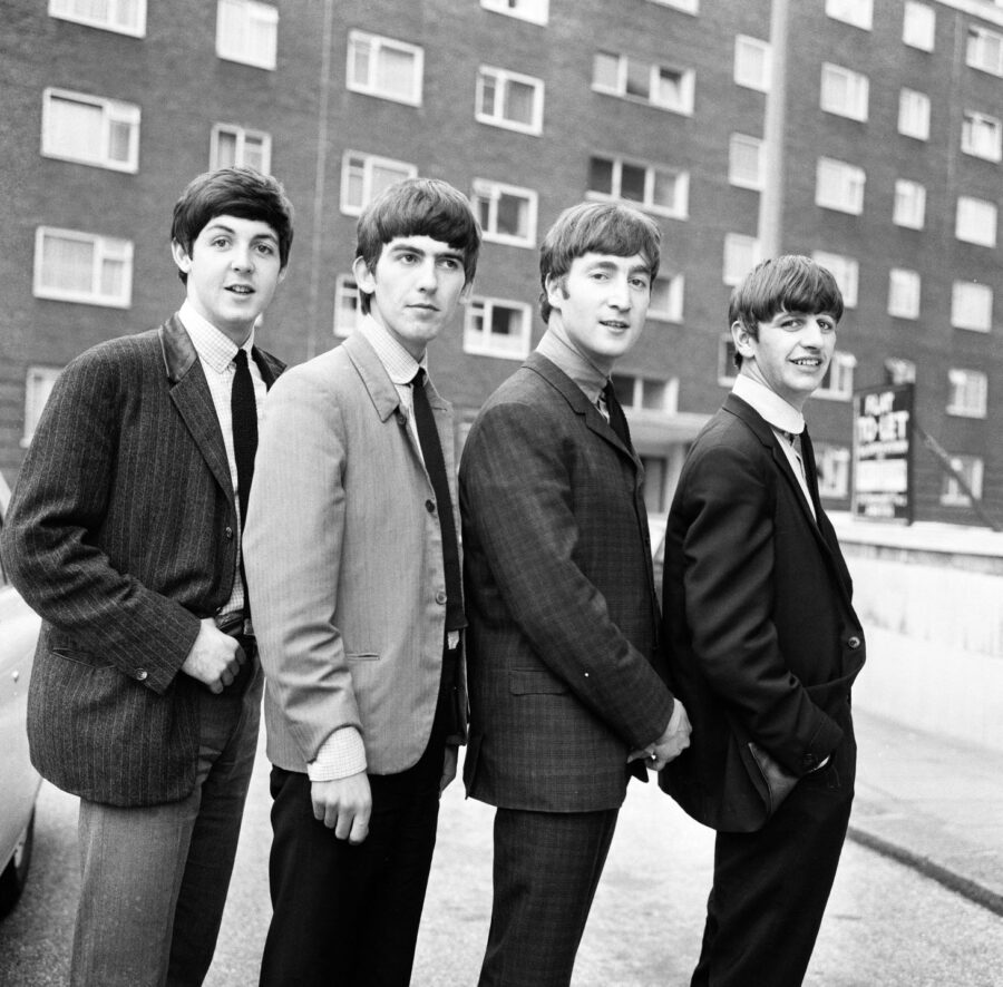 The Beatles pose in stylish suits against a brick building backdrop, showcasing their iconic early '60s fashion and charisma.