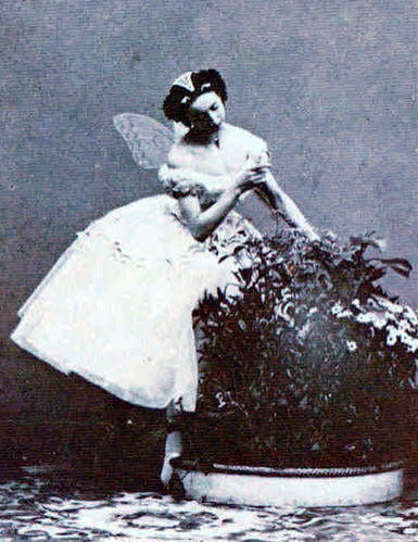 A young woman in a fairy costume, complete with wings, delicately tends to flowers in a large planter, capturing a whimsical, enchanting moment.