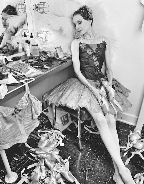 A dancer in a black and white setting rests at a cluttered dressing table, wearing a tutu, surrounded by ballet shoes and costumes, highlighting the behind-the-scenes of performance life.