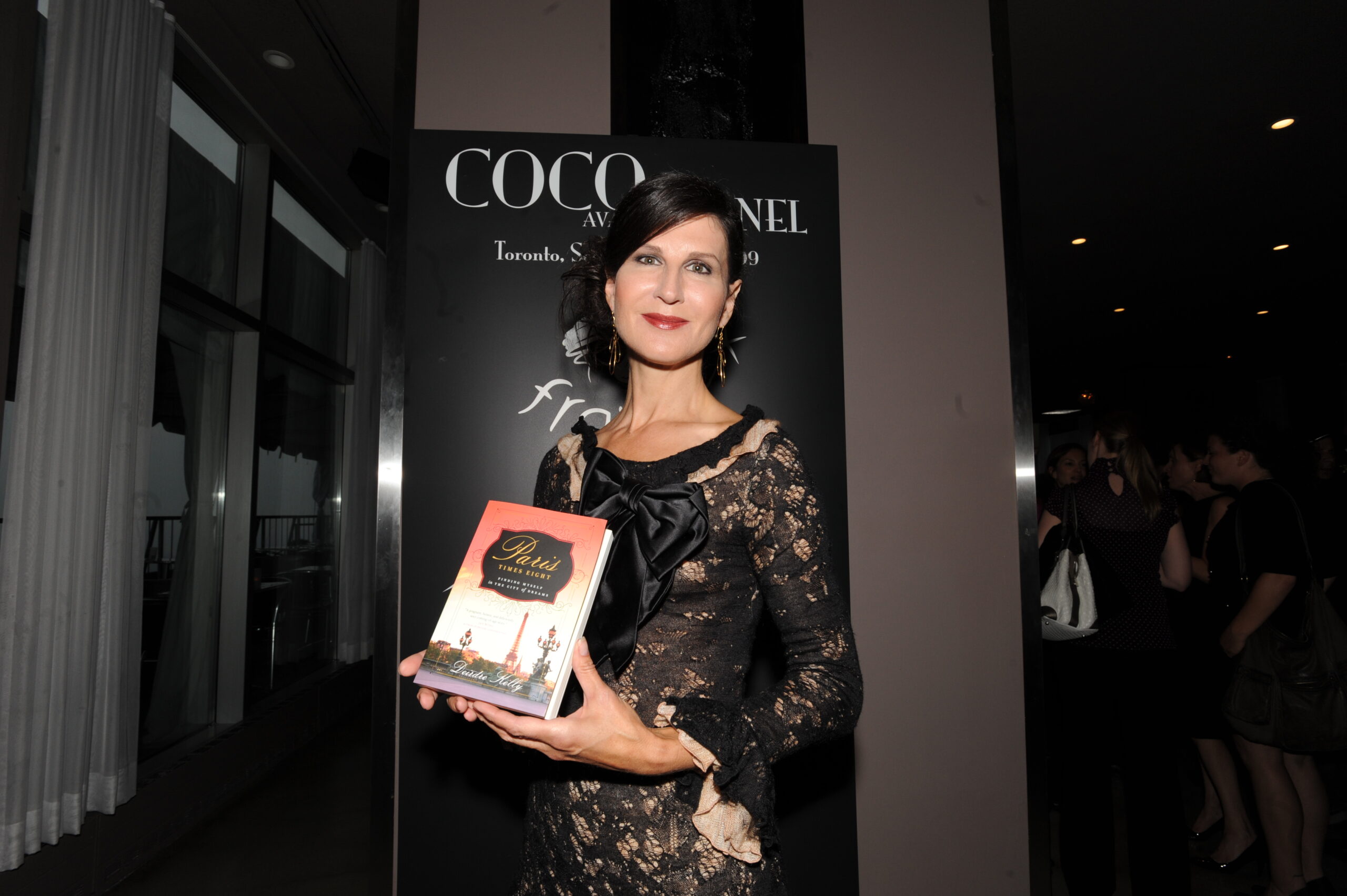 A woman in a lace dress holds a book titled "Paris: Times Eight" at a Coco Chanel event in Toronto, highlighting her literary work.