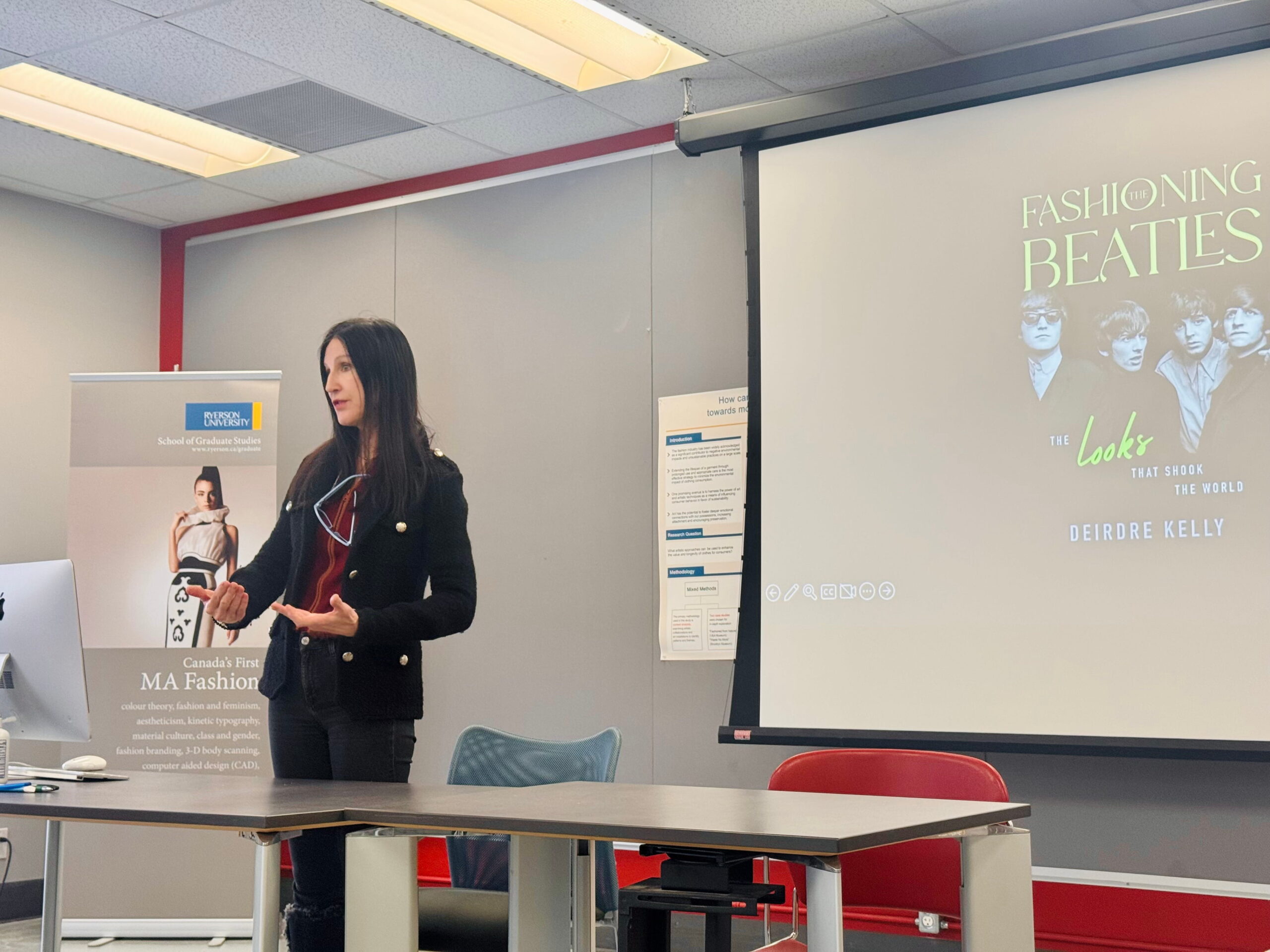 A woman presents in a classroom, gesturing towards a screen showing "Fashioning the Beatles: The Looks That Shook the World" by Deirdre Kelly. A banner beside her promotes a fashion program.