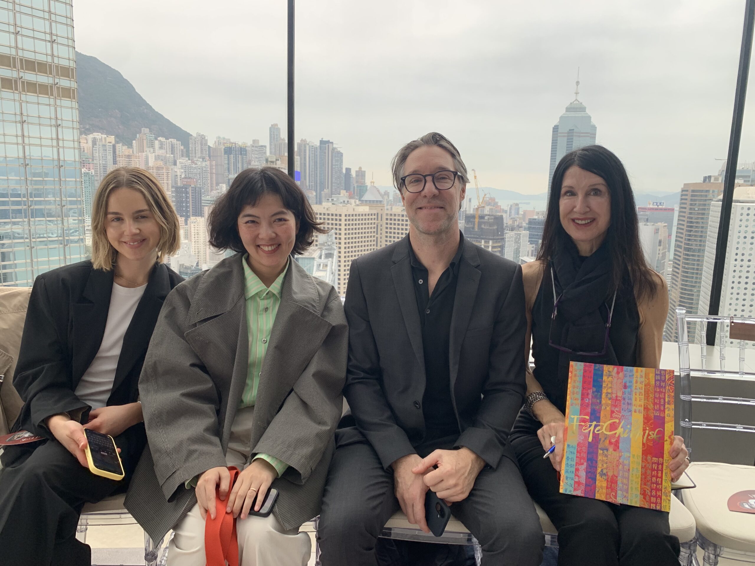 Four people seated on a rooftop with a cityscape background, smiling and dressed in dark clothing. The mood is upbeat and relaxed. One person holds a colorful book.